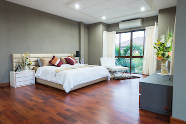 a larger room with a hardwood floor, a tall window overlooking trees, a two-person bed with a single bedside table, and a padded chair next to the window